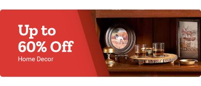 Promotional banner on a red background. Large white headline text reads: Up to sixty percent off. Smaller white subheadline text reads: Home Decor. On the right side of the banner is a rustic home decor display on a wooden shelf, including a framed deer illustration, a framed photo of a dog, a round wood slice tray holding small glass candle holders, and additional decorative accents arranged against a dark wood interior backdrop.