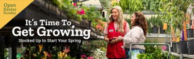 Open Easter Sunday. It is time to get growing. Stocked up to start your spring. A Tractor Supply Company employee wearing a red vest stands in a greenhouse aisle smiling and talking with a customer beside a shopping cart filled with plants, surrounded by shelves of colorful flowers and hanging greenery.