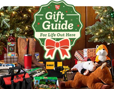 Holiday-themed promotional banner titled Gift Guide For Life Out Here featuring a green badge design with a red ribbon bow at the center. The background shows Christmas gifts under a decorated tree,including plush animals,toy trucks,and boxed toys. The warm lighting and rustic wooden floor create a cozy festive atmosphere highlighting holiday shopping ideas.